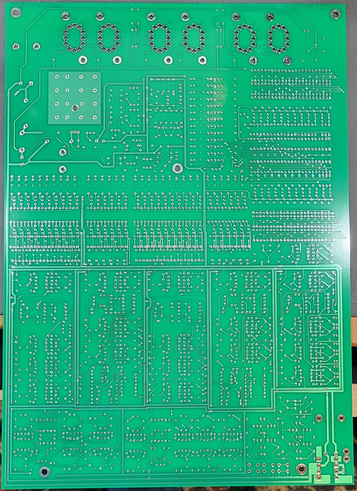 Figure 2 — The solder side: silkscreened "Version 5" at top left and "(c) KABtronics 2024" at top right, with the nixie wiring holes numbered 0–9 and COM below each socket. Photo: my board, 2026-…